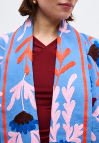 Γυναίκα που φοράει μπλε μαντίλι με ροζ και πορτοκαλί κεντήματα λουλουδιών πάνω σε μπορντό μπλουζάκι με V λαιμόκοψη, με εμφανή μόνο τα χείλη και το πηγούνι.