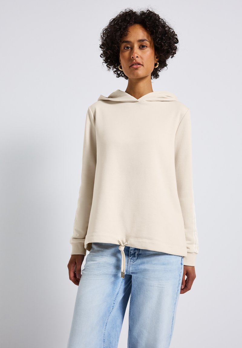 Felpa beige con cappuccio e cordino, caratterizzata da una vestibilità comoda e polsini a costine. Abbinata a jeans di denim azzurro chiaro.