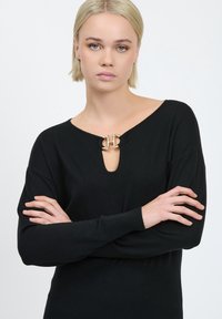 Schwarzer Strickpullover mit tiefem V-Ausschnitt, verziert mit einem goldenen Hardware-Akzent am Ausschnitt. Gerippte Bündchen und Saum sorgen für Textur.