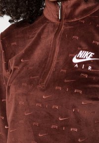 Osoba oblečená v hnědém Nike Air velurovém pulovru s polovičním zipem a vyraženými logy "Air" a swoosh po celém materiálu.