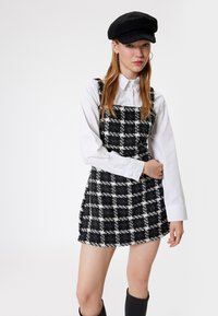 Vestido peto a cuadros en blanco y negro con tejido texturizado, escote cuadrado, superpuesto a una camisa blanca con botones; combinado con una gorra negra.