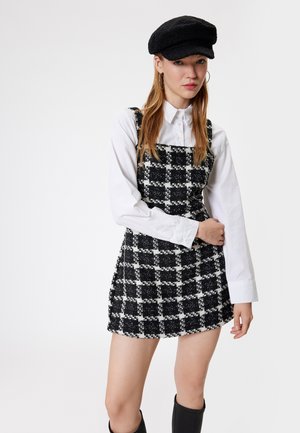 Abito a salopette a quadri bianco e nero con tessuto testurizzato, scollatura quadrata, indossato sopra una camicia bianca con bottoni; abbinato a un berretto nero.