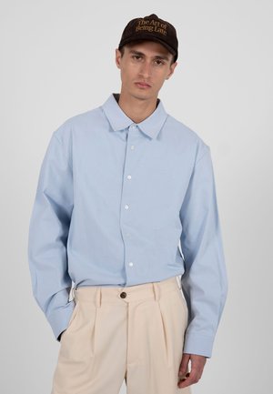 Mann mit brauner Kappe mit der Aufschrift "The Art of Being Late", hellblauem Hemd und beigen, plissierten Hosen vor einem einfarbigen Hintergrund.