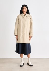 Beige trench coat with a collar, long sleeves, and button closure. Worn over a navy skirt, paired with cream socks and shoes. Flat, textured surface.
