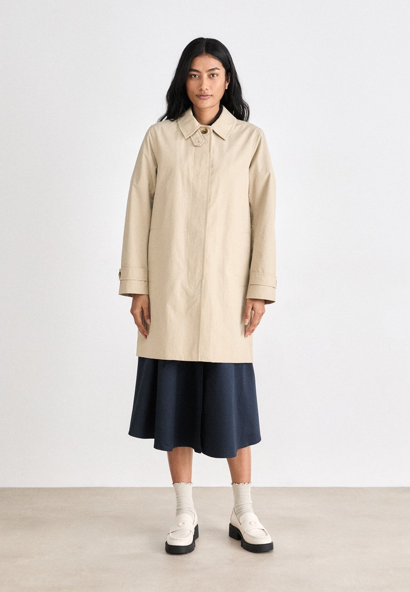 Gabardina beige con cuello, mangas largas y cierre de botones. Lleva una falda azul marino, acompañada de calcetas y zapatos crema. Superficie plana y texturizada.