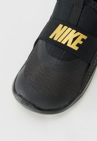 Svart Nike-sneaker med teksturert tå, mesh-øvre og bred stropp med gull "NIKE"-logo på hvit bakgrunn.