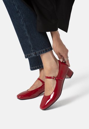 Fødder iført skinnende røde Mary Jane-sko med lav blokhæl, kombineret med mørkeblå jeans og en hånd, der justerer skospændet.