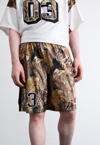 Camisola branca com o número "03" em preto e calções de camuflagem castanhos com uma cintura preta, detalhes em vermelho e um logótipo bordado na lateral.