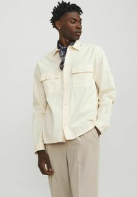 Chemise boutonnée beige clair avec deux poches poitrine, col standard et manches longues, associée à une écharpe à motifs bleu marine.