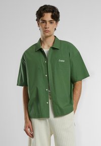 Camicia verde a maniche corte con colletto e tasca frontale, con testo ricamato bianco. Realizzata in tessuto morbido, con bottoni davanti.