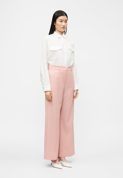 Γυναίκα που στέκεται φορώντας ένα λευκό πουκάμισο με μακριά μανίκια και κουμπιά, ροζ παντελόνια με φαρδιά πόδια και ψηλή μέση, και λευκές ψηλοτάκουνες γόβες με μυτερή μύτη.