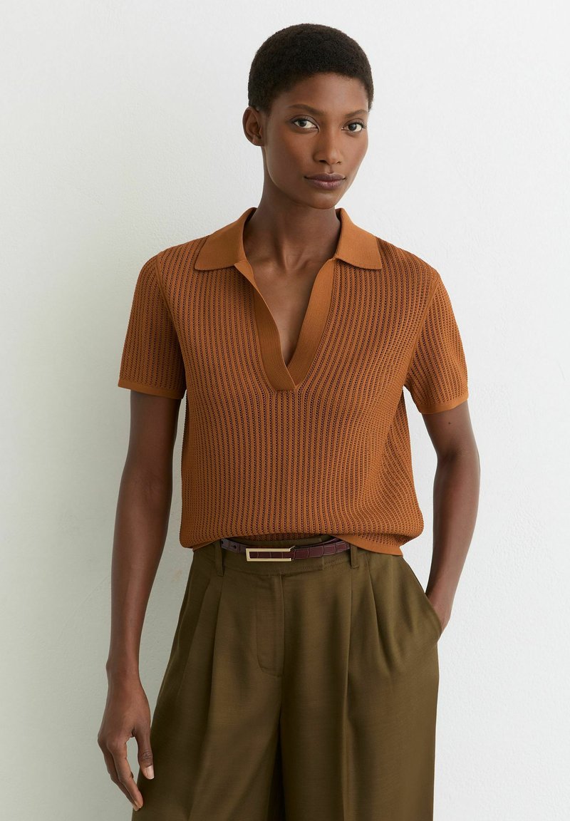 Nő, texturált rozsdabarna rövid ujjú felsőt visel, mély V-nyakú gallérral, valamint olívazöld magasderekú nadrágot barna övvel.