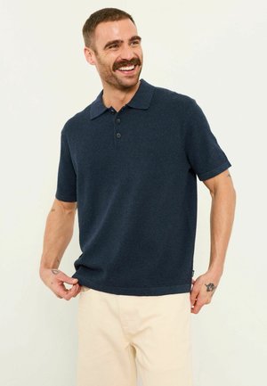 Lächelnder Mann mit kurzen Haaren und Schnurrbart, trägt ein marineblaues Strick-Poloshirt und beigen Hosen, hält den Saum des Shirts und zeigt Tätowierungen auf den Armen.