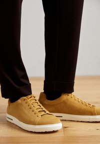 Gule ribbede sneakers med hvite såler, med rund tå, snøring og teksturert overflate, brukt med svarte oppbrettede bukser på tregulv.