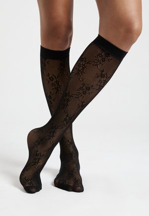 Calze nere in pizzo alte fino al ginocchio, caratterizzate da un motivo floreale, una texture in pizzo e una banda nera solida nella parte superiore. Design open toe.