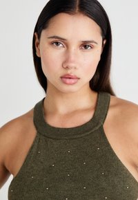 Top senza maniche di colore verde oliva con un collo alto, decorato con piccoli chiodini color oro rosato distribuiti uniformemente sul tessuto.