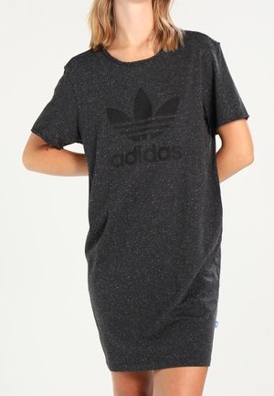 Femme portant une robe t-shirt Adidas gris foncé chiné avec logo, debout avec un bras derrière le dos sur un fond blanc.