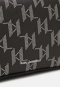 Musta tekstuuripintainen nahkalaukku, jossa on toistuva valkoisista pisteistä koostuva logo-kuvio. Metallinen logo-yksityiskohta laukun alaosassa. Kompaktin ja suorakaiteen muotoinen.