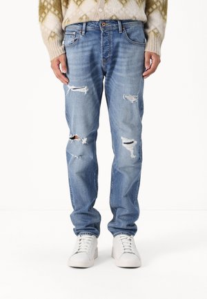 Jean bleu clair déchiré à plusieurs endroits, porté avec des baskets blanches et un pull beige à motifs, visible de la taille aux pieds.