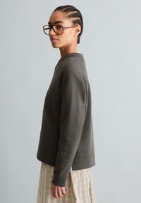 Junge Person mit cornrows und Brille trägt einen dunkelgrauen Pullover und einen karierten Rock, steht seitlich vor neutralem Hintergrund.