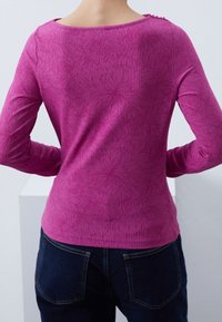 Langärmliges Shirt in leuchtendem Pink, mit strukturiertem Muster und Rundhalsausschnitt, kombiniert mit dunklen Jeans.