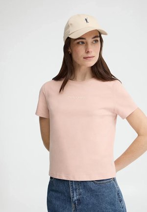 Mujer con camiseta rosa claro "Polo Club", gorra de béisbol beige con logo y vaqueros azules, mirando hacia un lado con las manos detrás de la espalda.