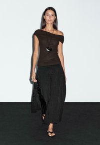 Schwarzes Off-Shoulder-Top kombiniert mit einem schwarzen, plissierten Maxirock. Das Model trägt eine große schwarze Anhänger-Halskette und schwarze Sandalen.