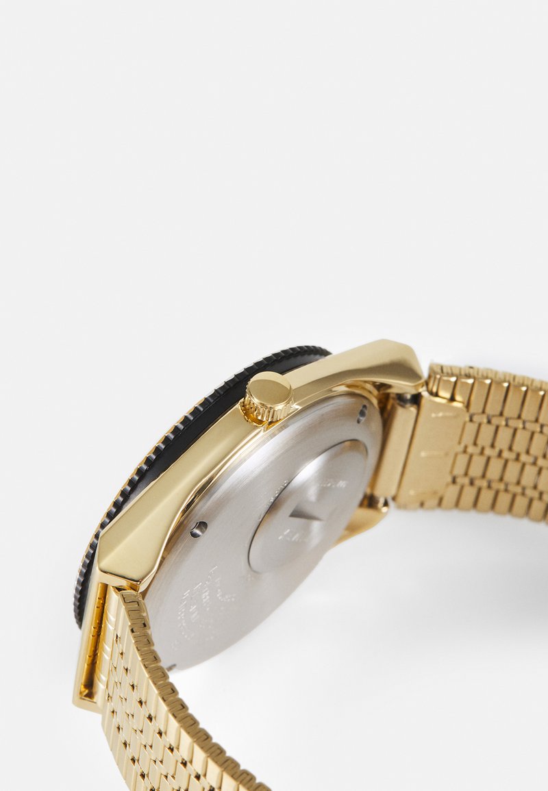 Reloj de metal en tono dorado con un bisel negro texturizado, caja redonda y pulsera trenzada. Presenta una corona prominente para el ajuste de la hora.