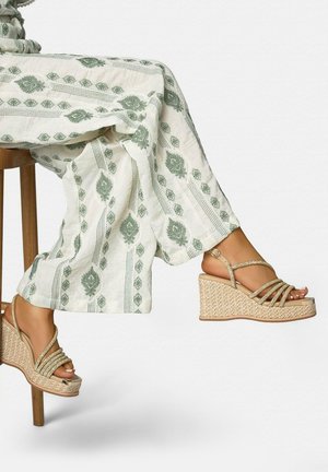Gambe con pantaloni bianchi con motivi verdi, sedute su uno sgabello di legno, e sandali beige intrecciati con zeppa e più cinturini.
