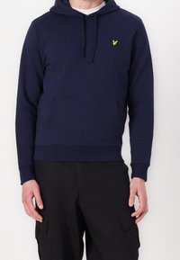 Homme portant un sweat à capuche bleu marine avec un logo jaune sur la poitrine et un pantalon noir avec des poches latérales, debout devant un fond uni.
