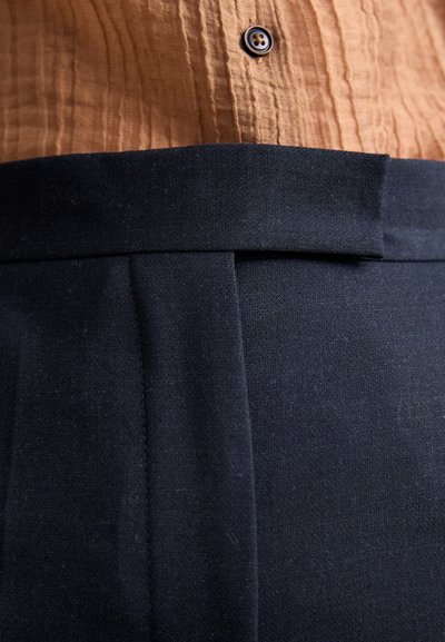 Sort, skræddersyede bukser med en struktureret talje, der har en plisseret front og en glat tekstur. Brun knapdetalje synlig ovenfor.