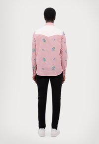 Rosa Hemd mit weißem Schulterbereich, verziert mit verstreutem blauen Blumenstickereien; kombiniert mit schwarzen Hosen und weißen Sneakers.