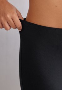 Zwarte leggings van zachte, rekbare stof, met een aansluitend ontwerp en een elastische tailleband, benadrukt door een paar handen die de taille verstellen.