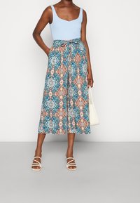 Un débardeur bleu associé à des culottes à motifs comportant des designs floraux bleus, oranges et blancs, avec une taille à liens. Sac à main blanc et sandales.