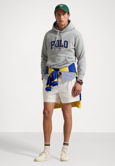 Šedá mikina s modrým logom "POLO", biele šortky, modro-žltý šál, zelená čiapka, biele tenisky a pruhované ponožky. Neformálny outfit s vrstvenými prvkami.