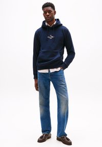 Felpa blu navy con tasca frontale e ricamo leggero, abbinata a jeans dritti blu e mocassini marrone scuro, in piedi contro uno sfondo bianco.