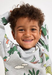 Lächelndes Kind mit lockigem Haar trägt einen hellgrauen Sweatshirt mit bunten Dinosaurier- und Wolkenmotiven, die Arme hinter dem Kopf erhoben.