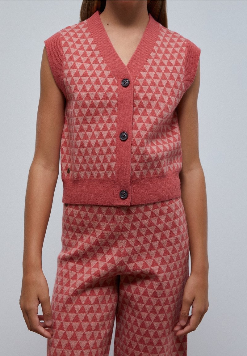 Gilet sans manches à boutons et pantalon assorti avec un motif de triangles géométriques rose et beige, porté par une personne aux bras détendus.