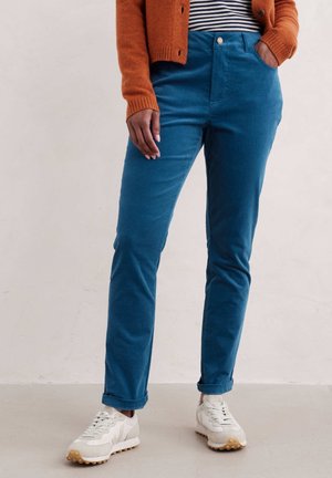 Person iført blå slim-fit bukser, orange cardigan, stribet skjorte og hvide sneakers, stående med den ene hånd i lommen.