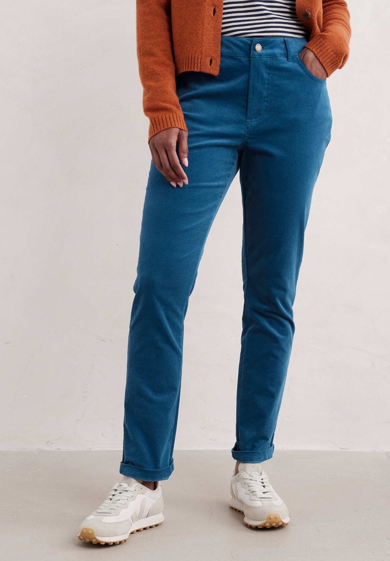 Personne portant un pantalon slim bleu, un cardigan orange, une chemise rayée et des baskets blanches, debout avec une main dans la poche.