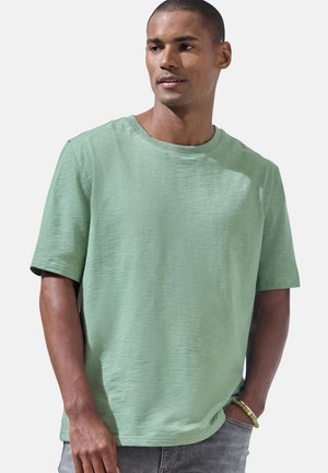 Mann trägt ein hellgrünes T-Shirt mit kurzen Ärmeln und graue Hose, schaut nach rechts, hat eine Hand in der Tasche und ein Perlenarmband am Handgelenk.