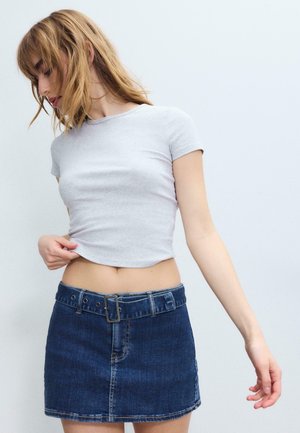 Jonge vrouw met blond haar, gekleed in een lichtgrijs cropped T-shirt en een blauwe denim minirok met een riem, staand tegen een effen achtergrond.
