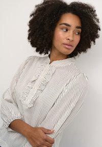 Blouse blanche avec des rayures noires verticales, un plastron froncé à l'avant et des poignets élastiques avec de longues manches. Tissu doux et coupe décontractée.