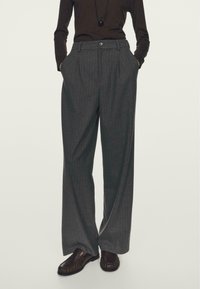 Grijze pinstripe broek met een hoge taille, wijde benen, manchetten aan de voorkant en zijzakken. Vergezeld van bruine schoenen.