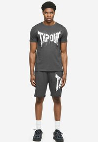 Man die staat en een zwart Tapout T-shirt draagt met bijpassende korte broek, witte sportsokken en zwarte sportschoenen op een witte achtergrond.