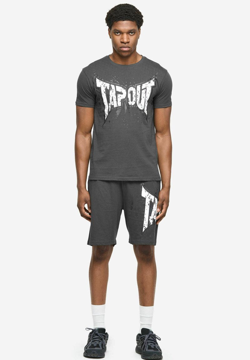 Man die staat en een zwart Tapout T-shirt draagt met bijpassende korte broek, witte sportsokken en zwarte sportschoenen op een witte achtergrond.