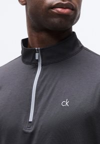 Κοντινό πλάνο ενός ατόμου που φοράει σκούρο γκρι τοπ με φερμουάρ και λευκό φερμουάρ και λευκό λογότυπο "ck" στο στήθος.