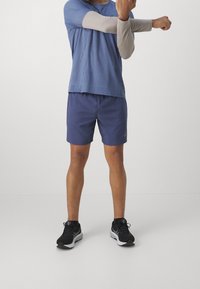 Man i blå atletiska shorts och en blå kortärmad t-shirt över en grå långärmad t-shirt, med svarta löparskor som sträcker ut en arm över bröstet.