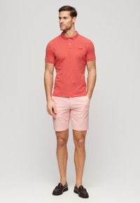 Camisa polo roja de manga corta, pantalones cortos rosas y mocasines negros. La camisa presenta un pequeño logo en el pecho y un diseño de cuello clásico.