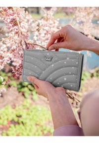 Gray quilted wallet with pink splatter, featuring a silver zipper and logo, held in a hand near blossoming flowers and green foliage.